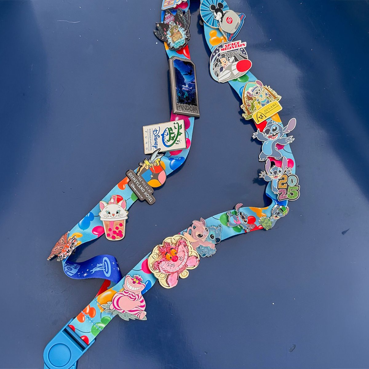 A colorful lanyard covered with various pins featuring cartoon characters and designs, including a Cheshire Cat, a frog, and other playful motifs, is displayed on a dark blue surface.