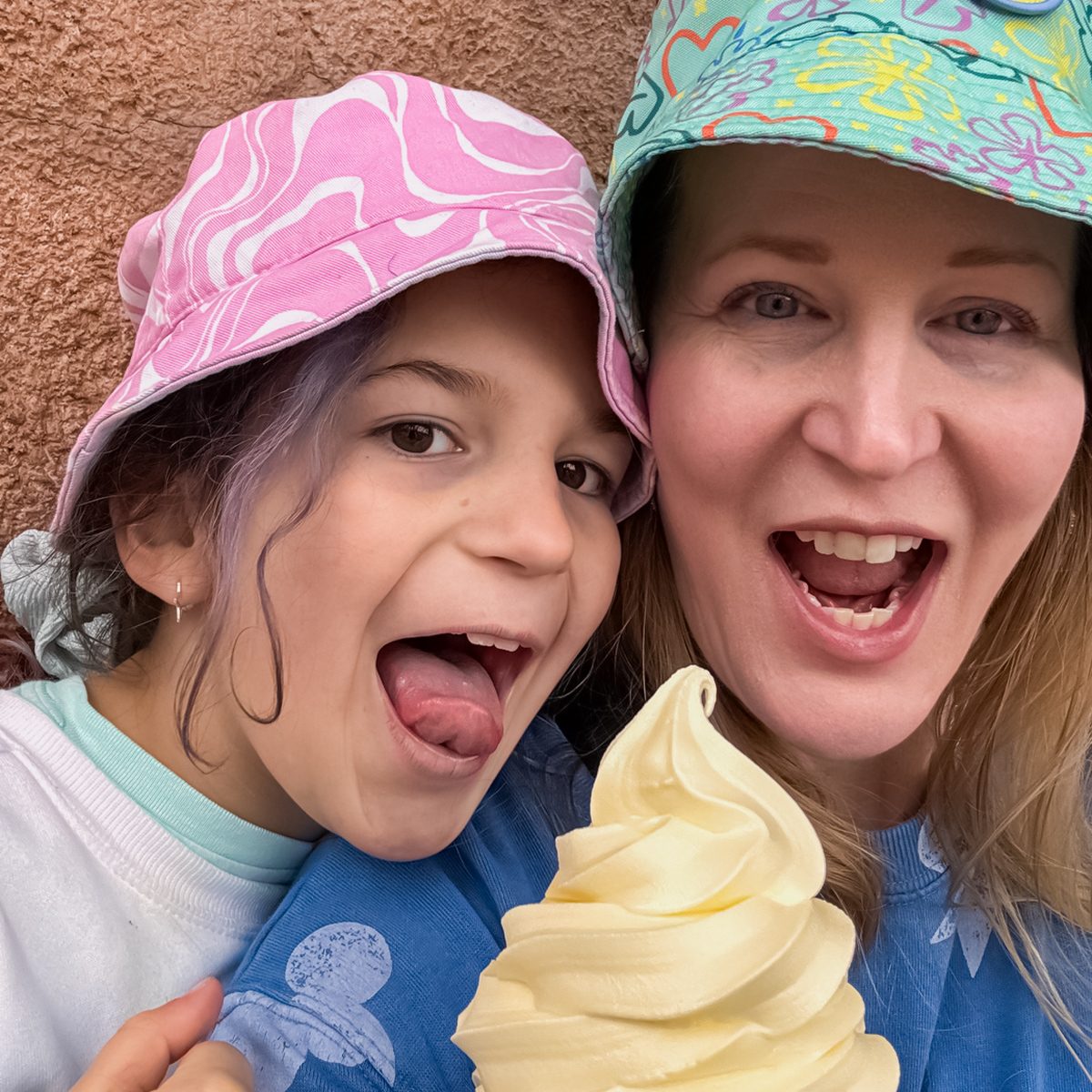 A woman and a young girl wearing colorful hats smile at the camera. They are holding a large swirl of soft-serve ice cream. The girl playfully sticks out her tongue towards the ice cream.