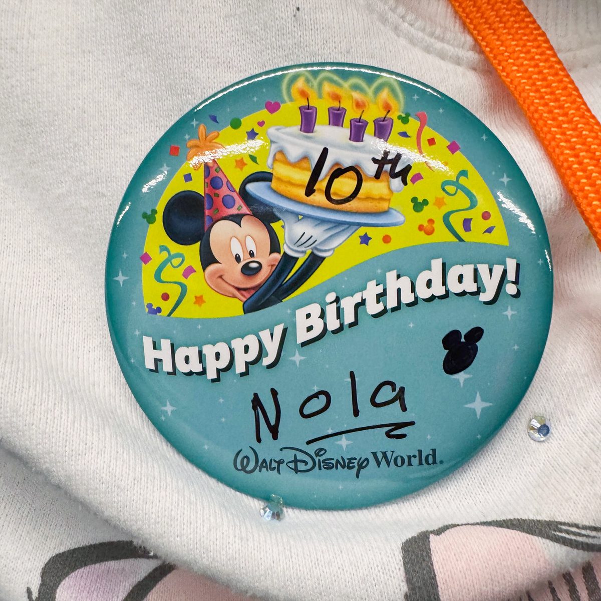 A Disney-themed birthday button with Mickey Mouse holding a cake adorned with candles. The button reads "Happy Birthday!" and has "10th" written on the cake. The name "Nola" appears beneath.