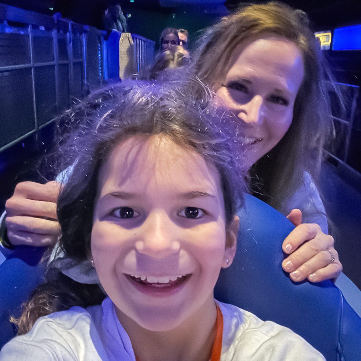 A girl and a woman smiling while seated, possibly on an amusement park ride. The background is dimly lit, suggesting an indoor setting with some lights in the distance.